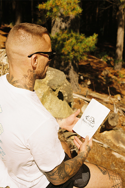 HCW A tattooed person in sunglasses and a white T-shirt sits outdoors in a peaceful forest, holding a white bag labeled HC Chocolates Extra Strength with a cloud illustration, surrounded by trees and rocks. Happy Cloud Wellness