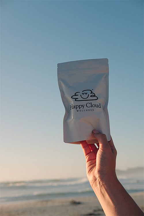 HCW A hand holds a white pouch labeled HC Chocolates Extra Strength with a smiling cloud logo, set against a clear blue sky and beach at sunset. Happy Cloud Wellness